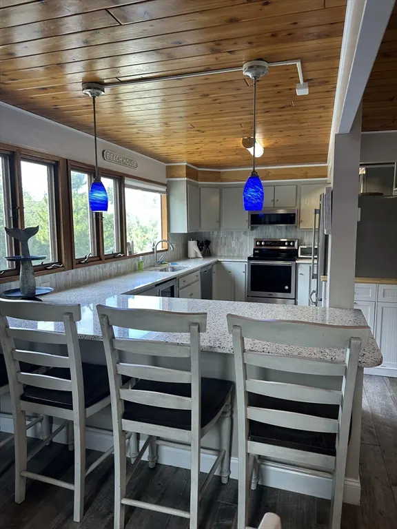 A bright kitchen with a wood ceiling, granite countertops, and blue pendant lights over a breakfast bar with white chairs.