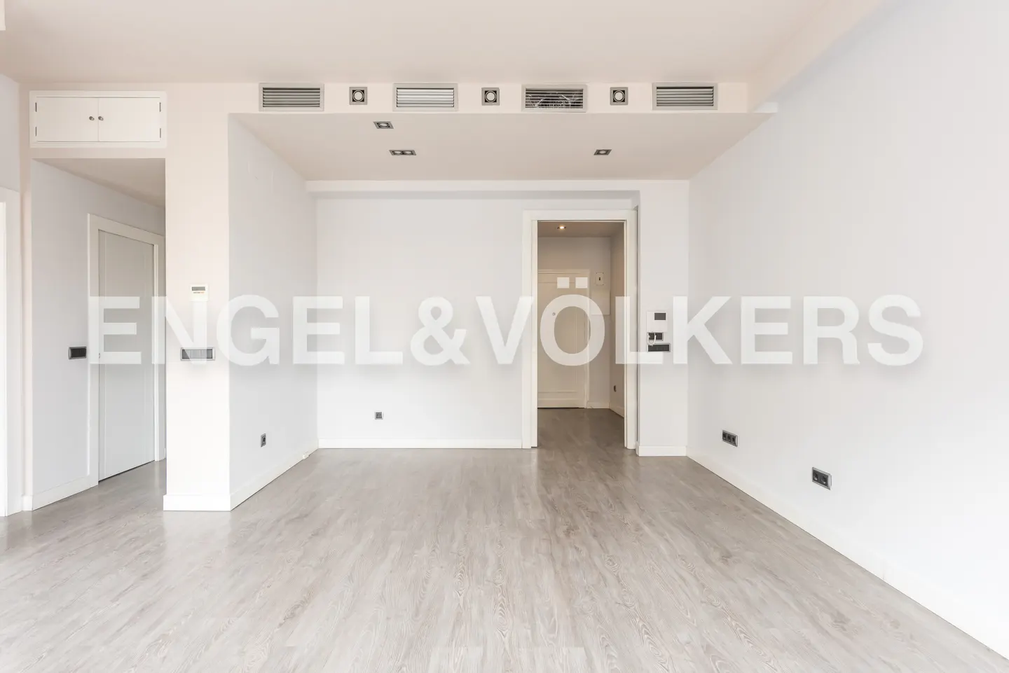 Empty, minimalist room with white walls, light wood-look flooring, recessed ceiling lights, and a doorway to another room.