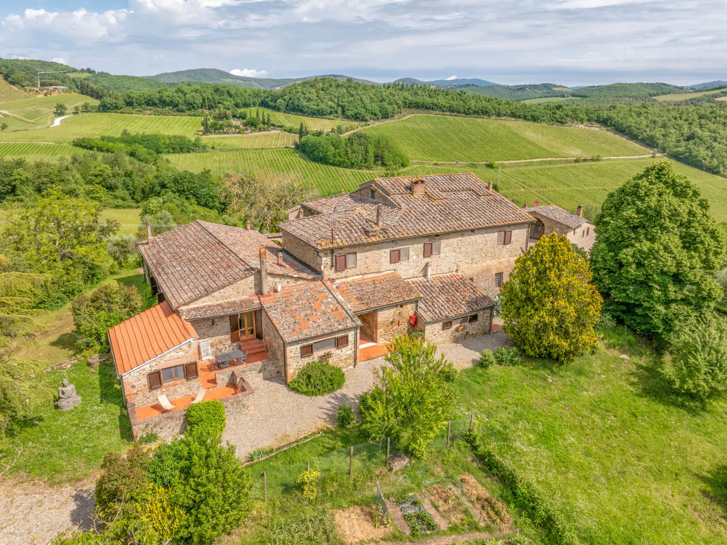 Aerial view of a stone farmhouse with orange tile roof, surrounded by green fields, trees, and vineyards.
