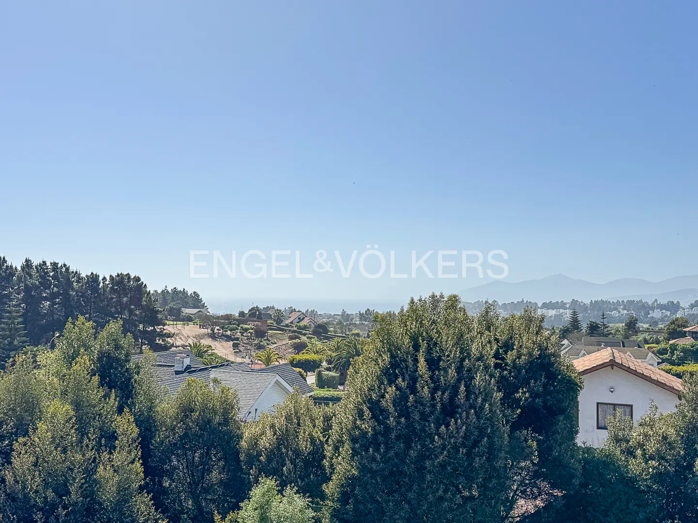 Real estate view of houses and trees under a blue sky, with mountains in the distance. Engel & Völkers logo in the center.