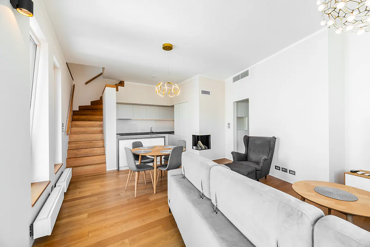 Bright, modern living space with wood floors, white walls, and a staircase. Features a gray sofa, dining table, and a sleek kitchen in the background.