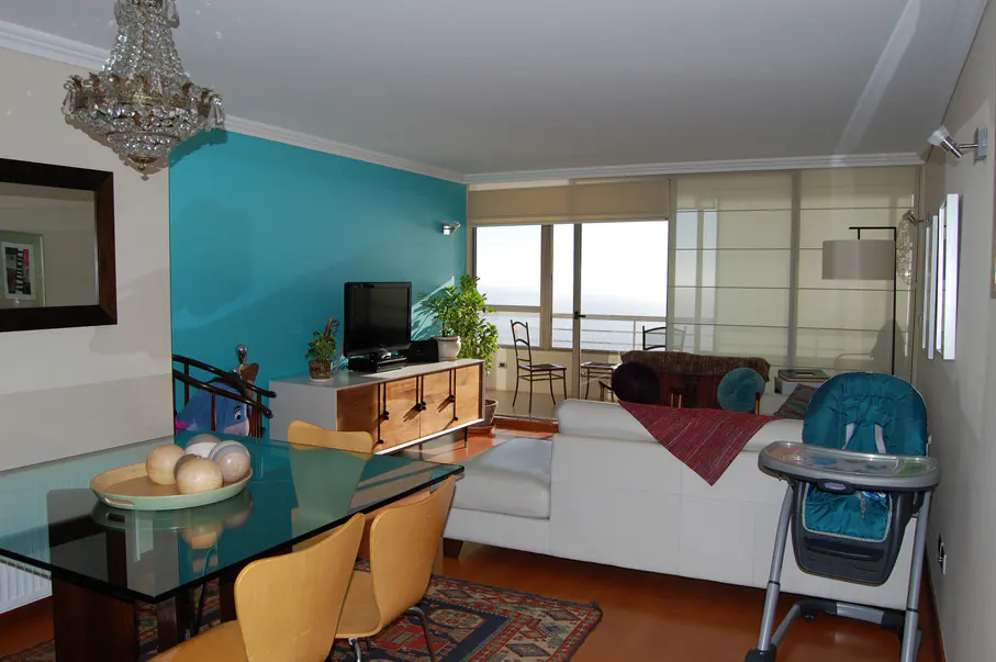 Bright living room with a glass table, white sofa, and a turquoise accent wall. A chandelier hangs above the table. Ocean view through the window.