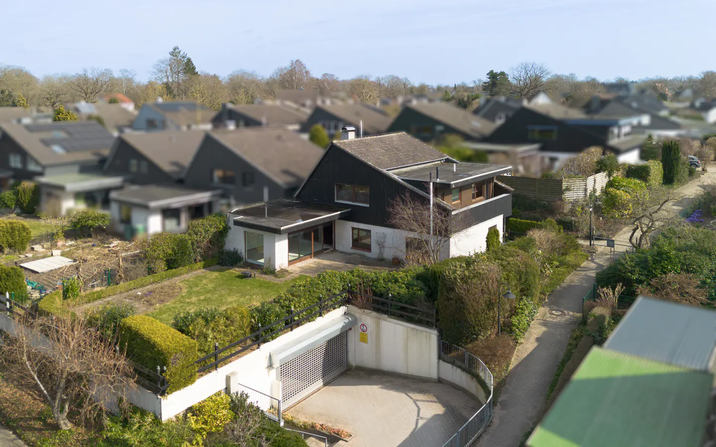 Aerial view of a two-story house with a black roof, white walls, and a garage entrance surrounded by green bushes.