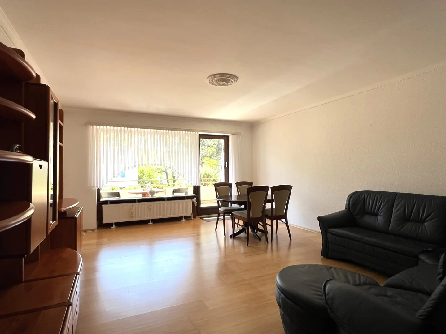 A bright living room with wood floors, a black leather sofa, a dining table with six chairs, and a large wooden cabinet.
