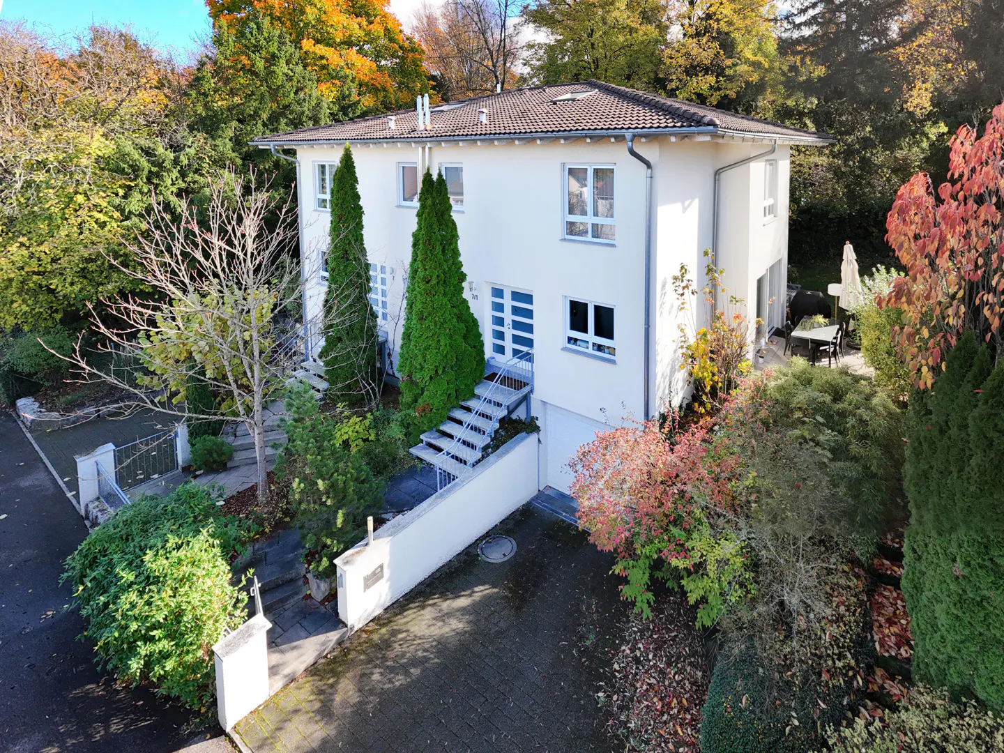 Two-story white house with a brown roof, surrounded by trees with autumn colors. A stone driveway leads to the house.