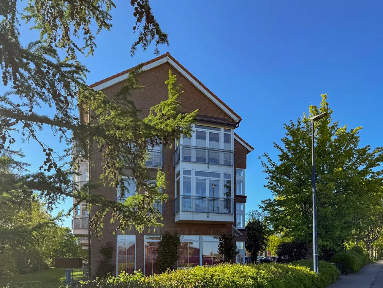 A three-story brick building with white trim and balconies, framed by green trees against a clear blue sky.