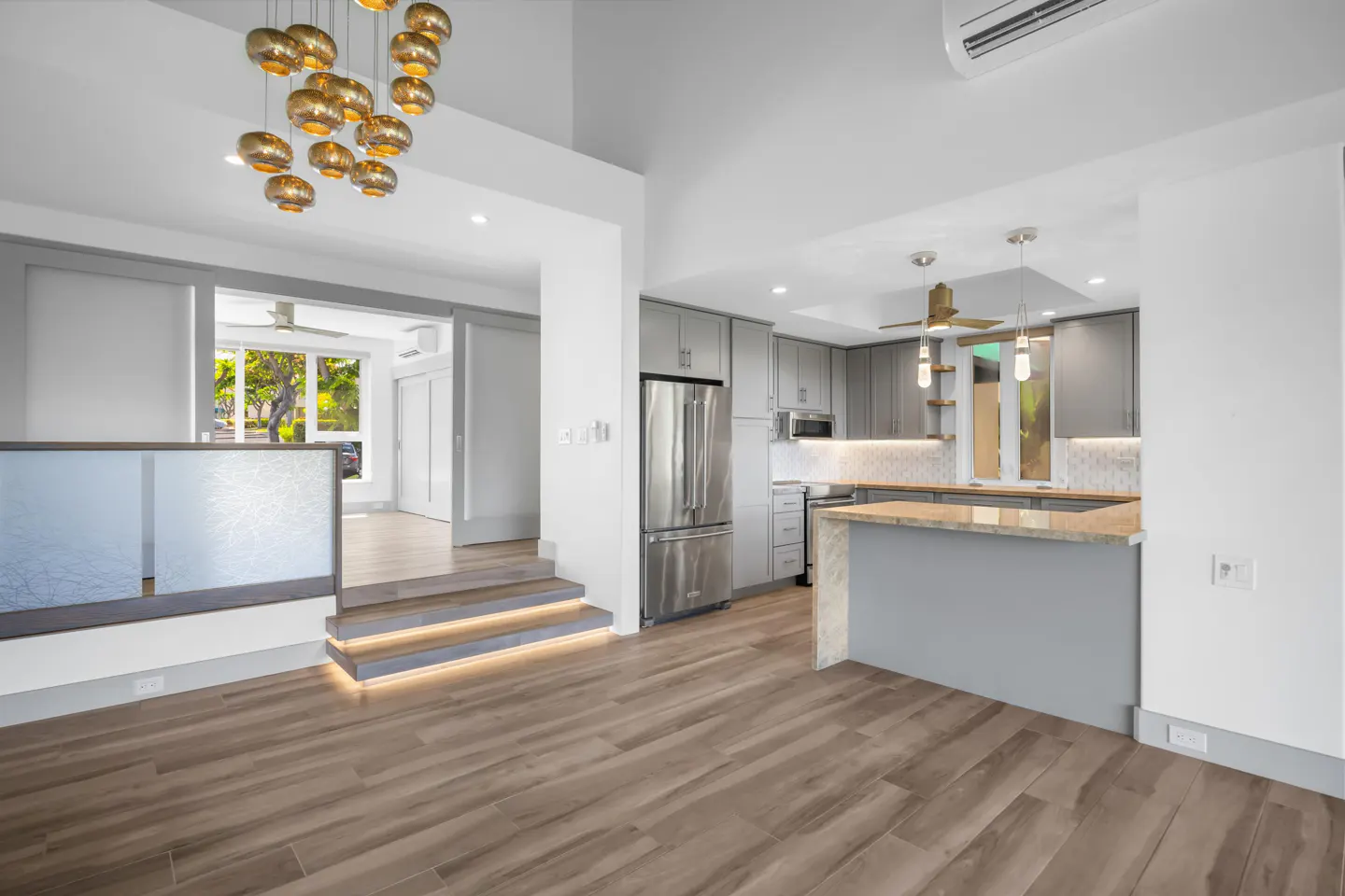 Modern kitchen with grey cabinets, stainless steel appliances, and a breakfast bar, featuring wood floors and gold lighting.