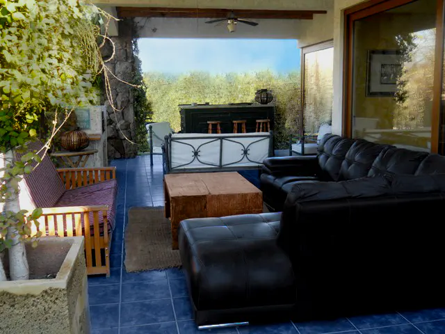 Outdoor living space with blue tile floor, black leather sofa, wood table, and bar area overlooking green trees.
