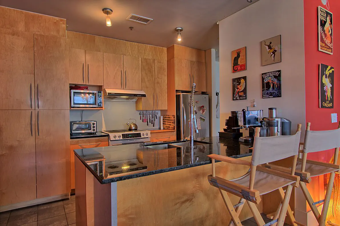 A modern kitchen with light wood cabinets, black granite countertops, and director's chairs at the island. Vintage posters decorate the walls.