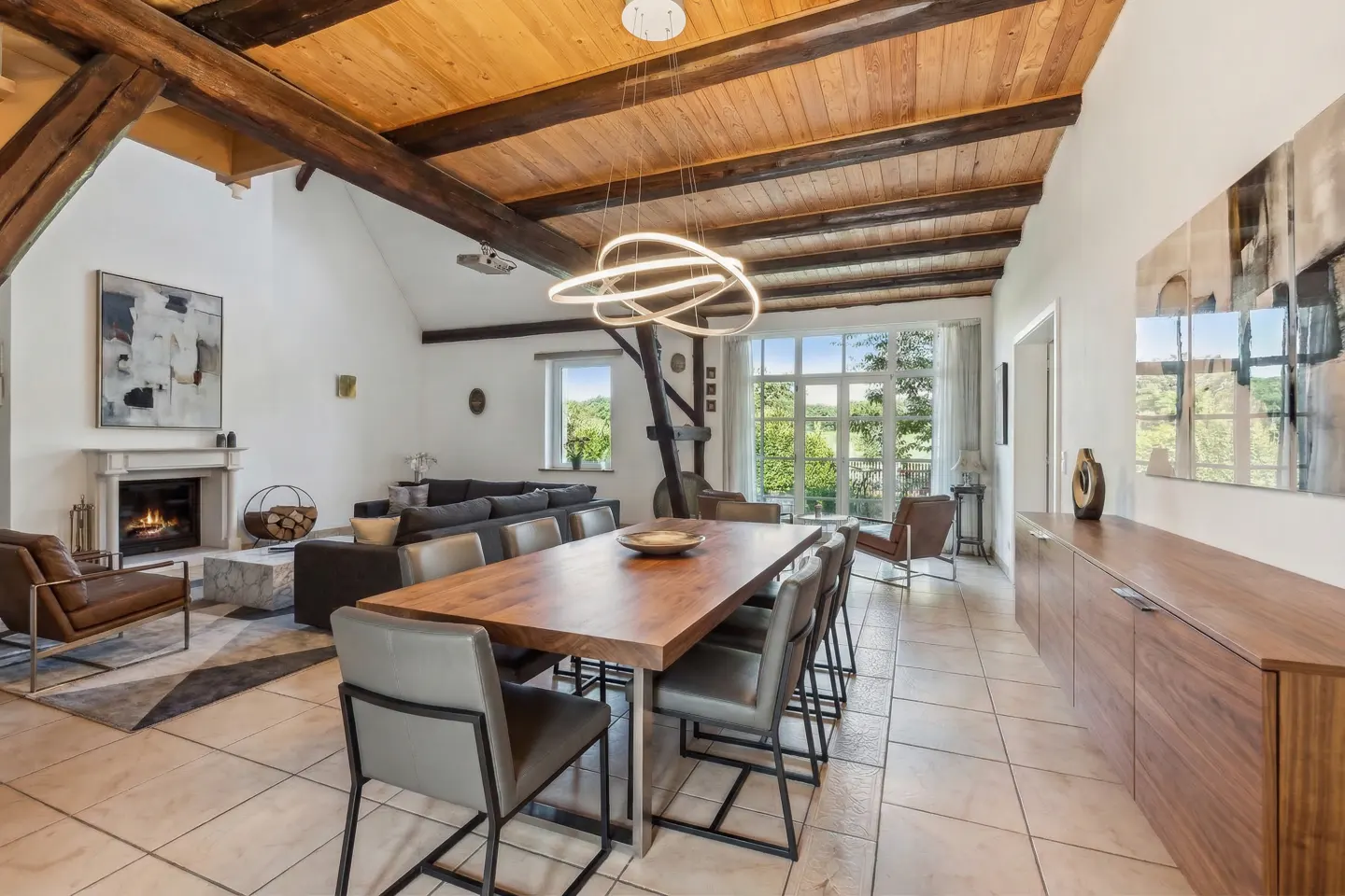 Open-concept living and dining area with wood ceiling beams, fireplace, and large windows overlooking greenery.