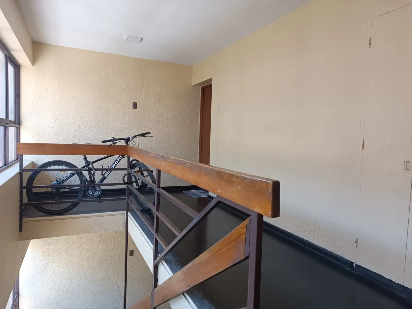 Interior view of a landing with a black bicycle parked behind a wooden and metal railing. Cream-colored walls and a black floor are visible.