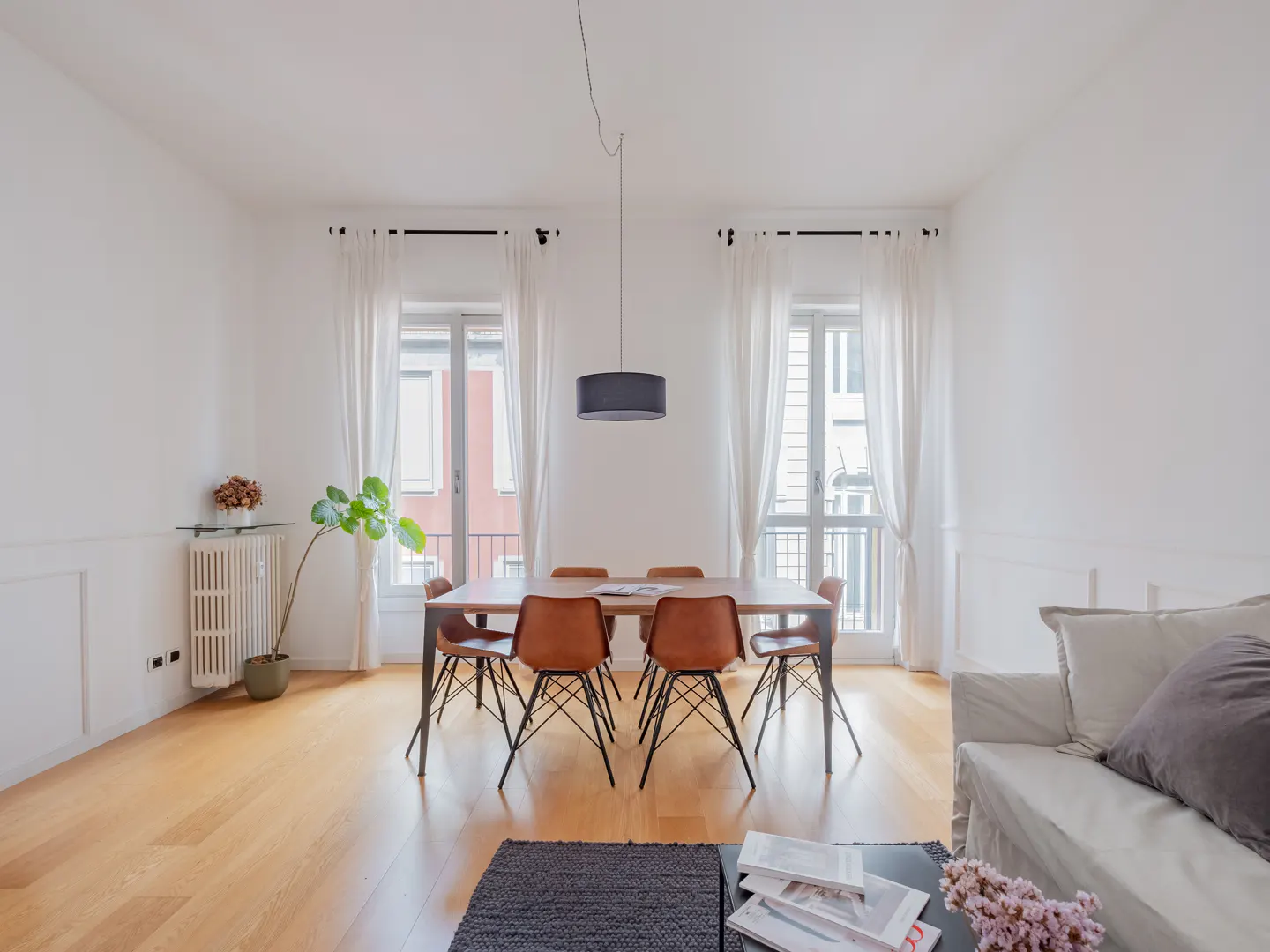 Bright living room with wood floors, white walls, and a dining table with six brown chairs. Two windows with white curtains flank the table. A sofa is visible in the foreground.