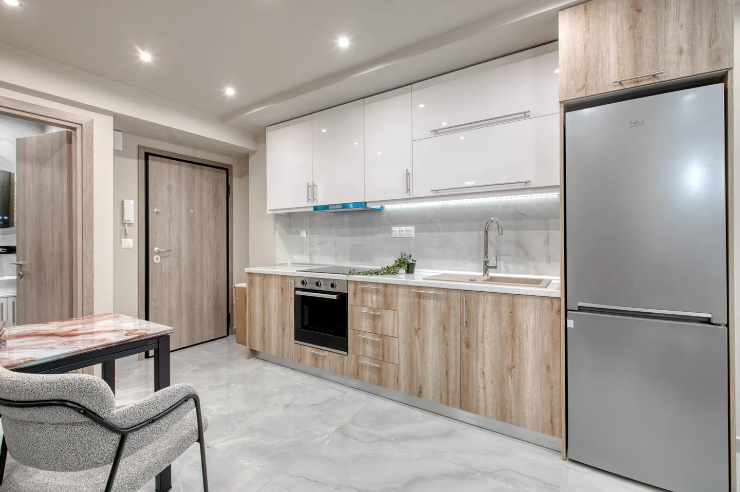 Bright kitchen with white cabinets, wood-grain lower cabinets, and a stainless steel refrigerator. A table and chair are in the foreground.