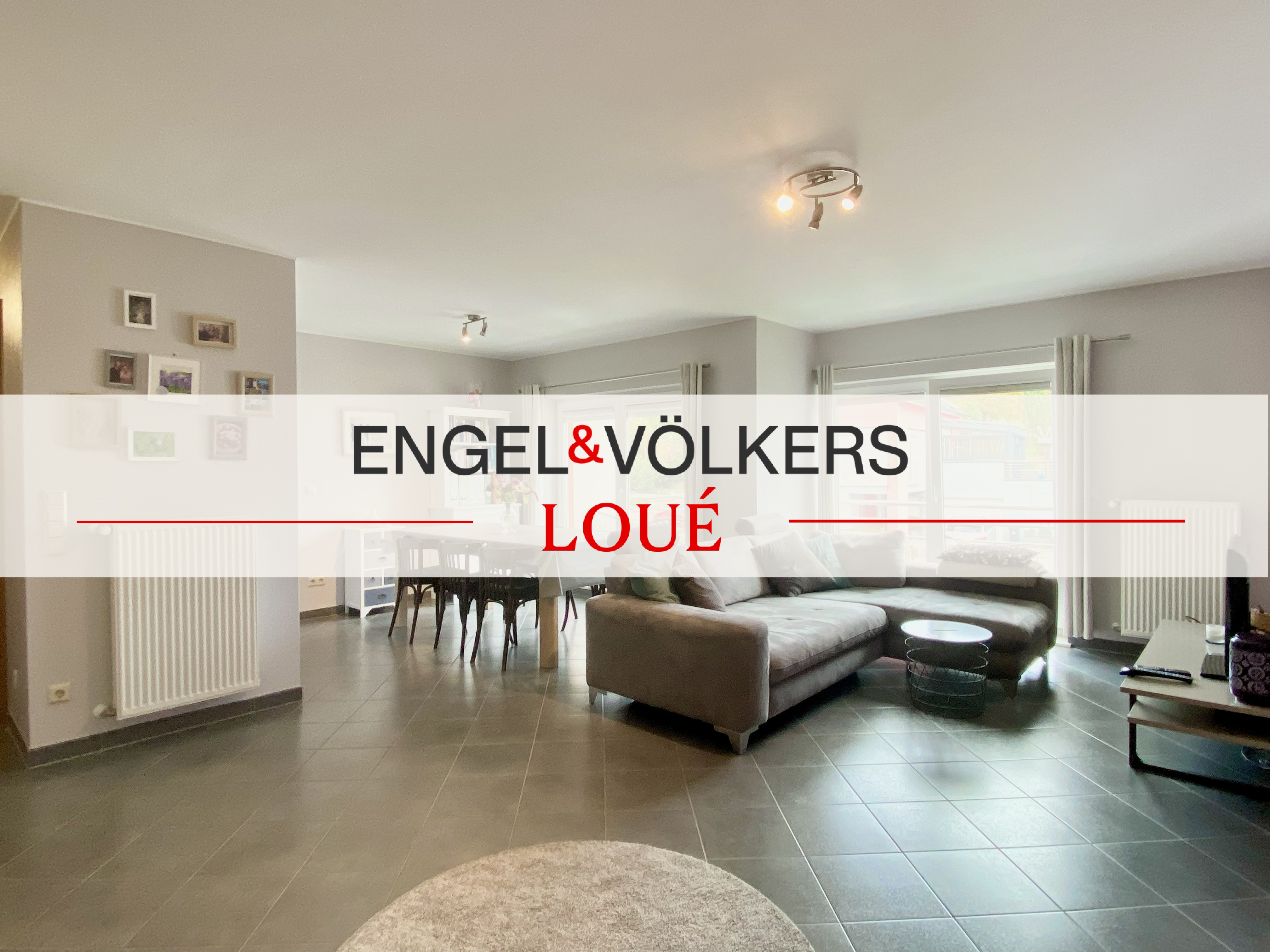 A wide shot of a living room with a gray sofa, dining table, and gray tiled floor. The Engel & Völkers logo is overlaid.