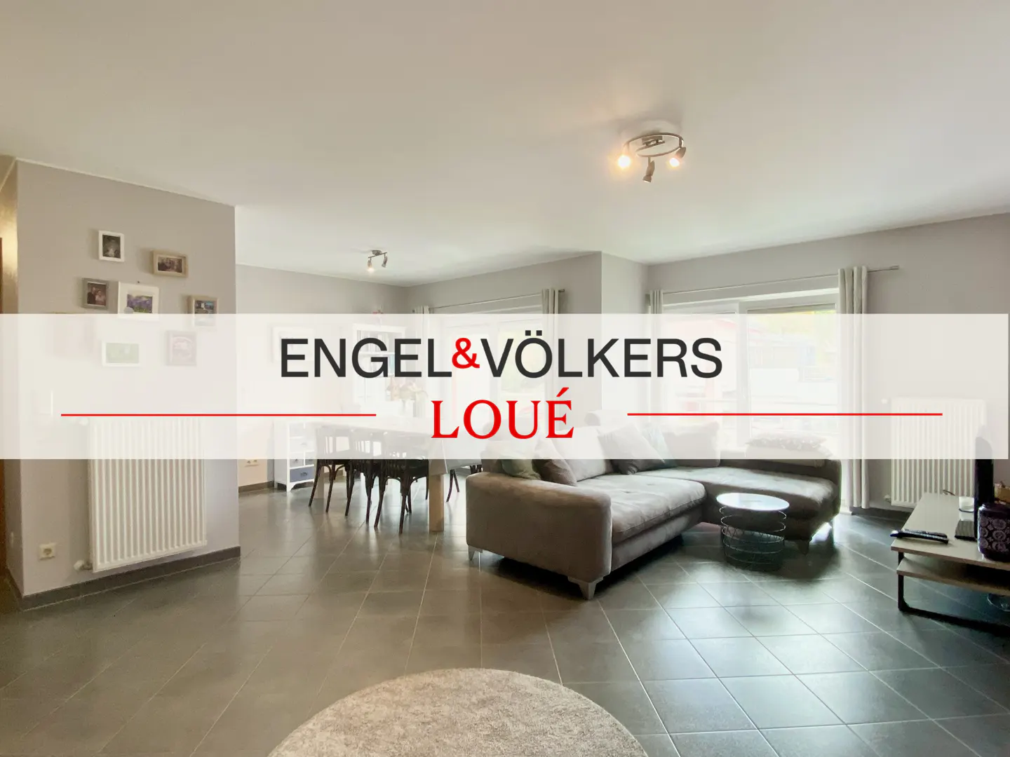 A wide shot of a living room with a gray sofa, dining table, and gray tiled floor. The Engel & Völkers logo is overlaid.