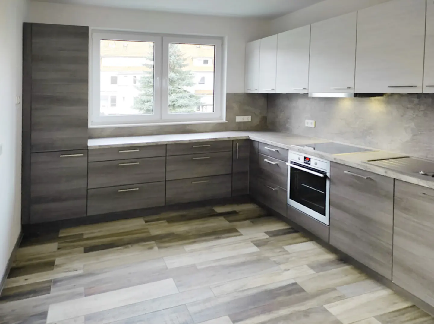 A modern kitchen with gray wood-look cabinets, white upper cabinets, and wood-look tile flooring. A window is above the counter.