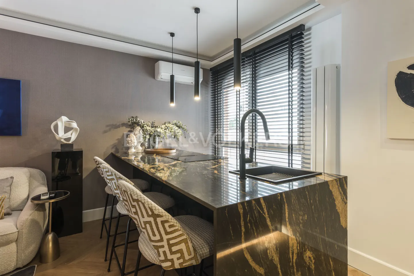 Modern kitchen island with black marble countertop, black faucet, and three pendant lights. Bar stools with patterned fabric. Window with blinds.