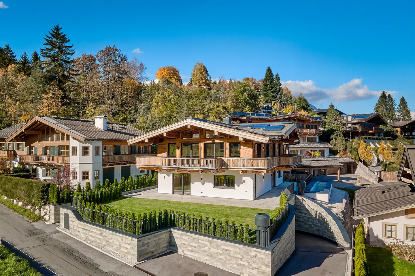 A chalet-style home with wood trim and a balcony sits on a green lawn, surrounded by trees and other houses on a sunny day.