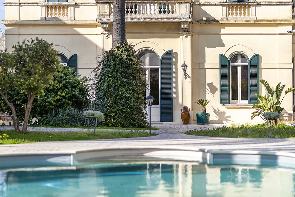 Exterior view of a cream-colored villa with green shutters, lush greenery, and a turquoise pool in the foreground.