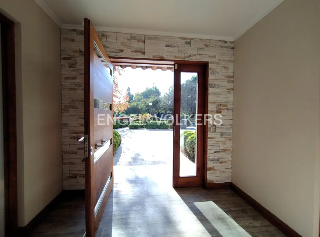 Open wooden front door with glass panels, stone wall, and view of a driveway and garden.