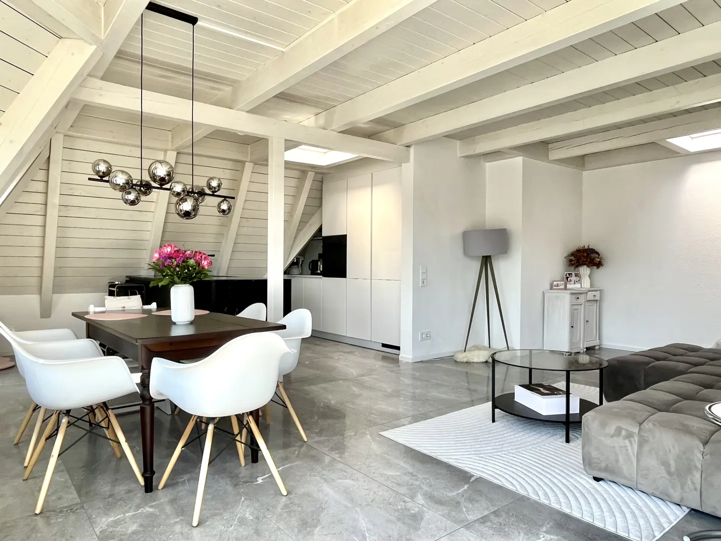 Bright, modern living space with white walls, exposed beams, and gray tile floors. A dining table with white chairs sits near a gray sofa.