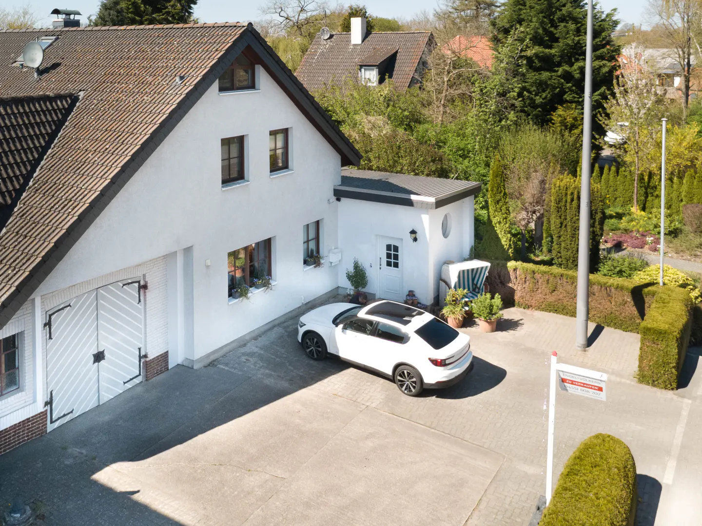 Aerial view of a white house with a brown roof, a white car parked in the driveway, and a "For Sale" sign in the yard.