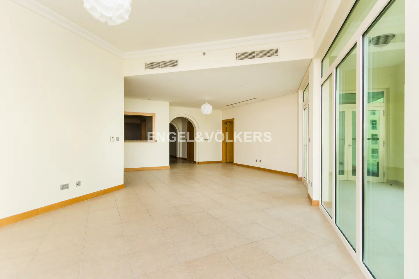 Empty, bright apartment with beige tile floors, white walls, and large windows. Archways lead to other rooms. A white light fixture hangs from the ceiling.