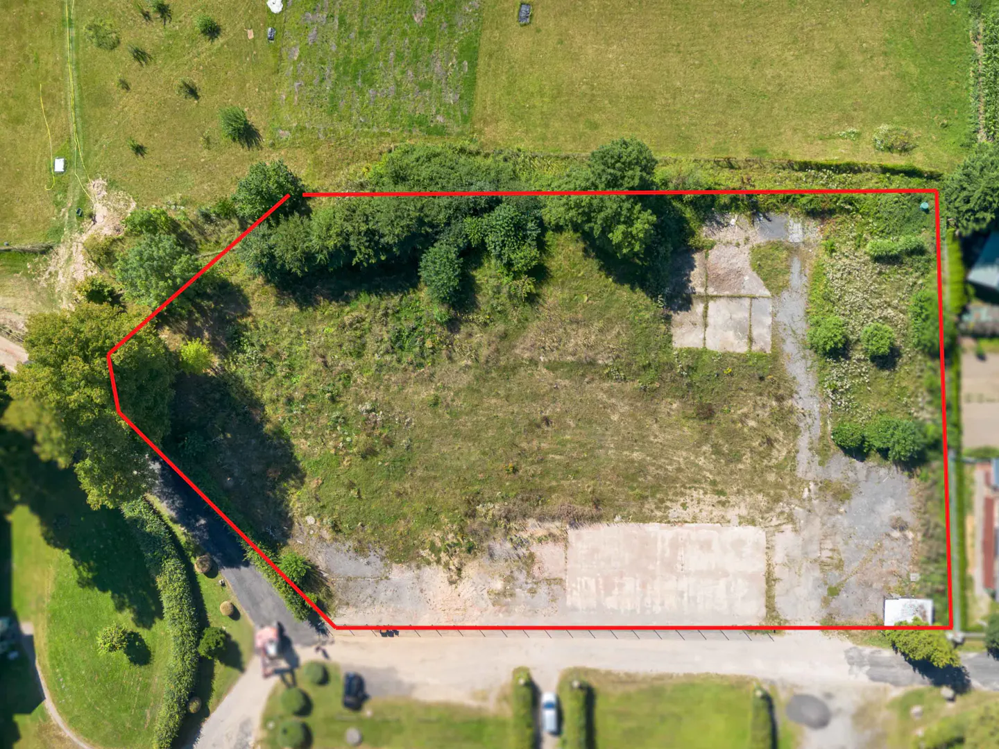 Aerial view of a vacant lot with a red border, surrounded by green trees and grass. Concrete slabs are visible on the lot.