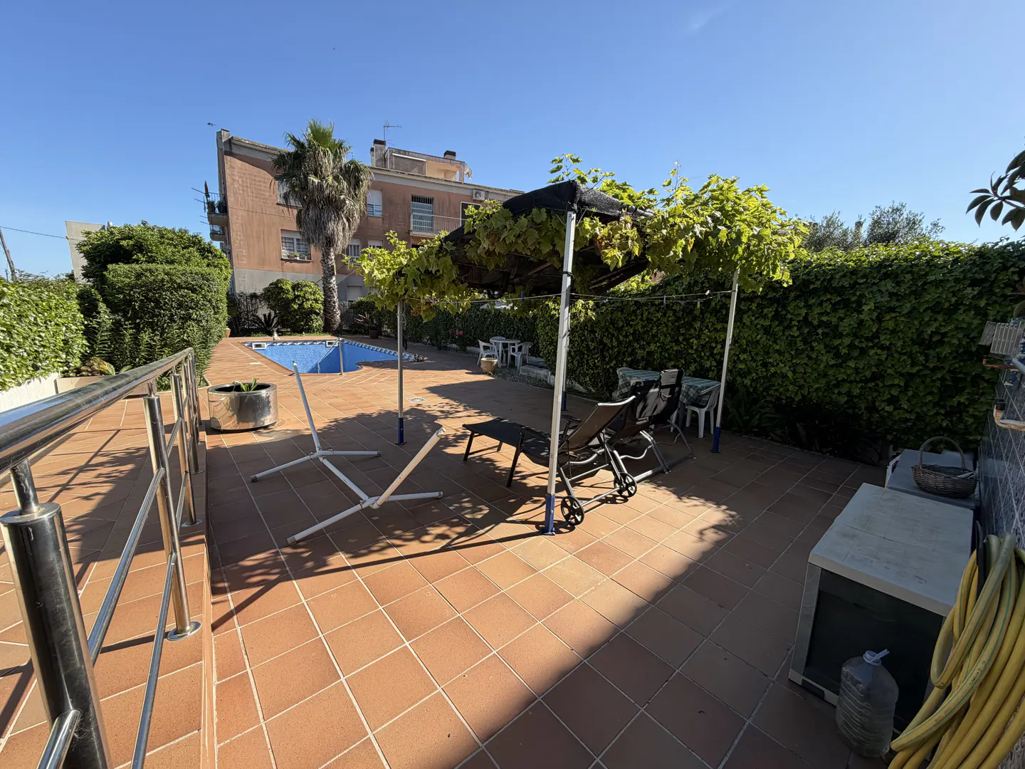 Outdoor patio with terracotta tiles, lounge chairs under a vine-covered pergola, and a blue pool in the background.