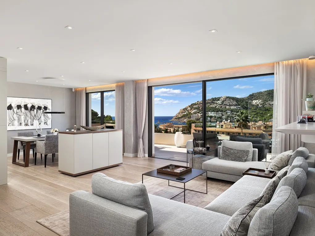 Bright, open-plan living room with a gray sectional sofa, wood floors, and a view of the ocean and mountains through large sliding glass doors.