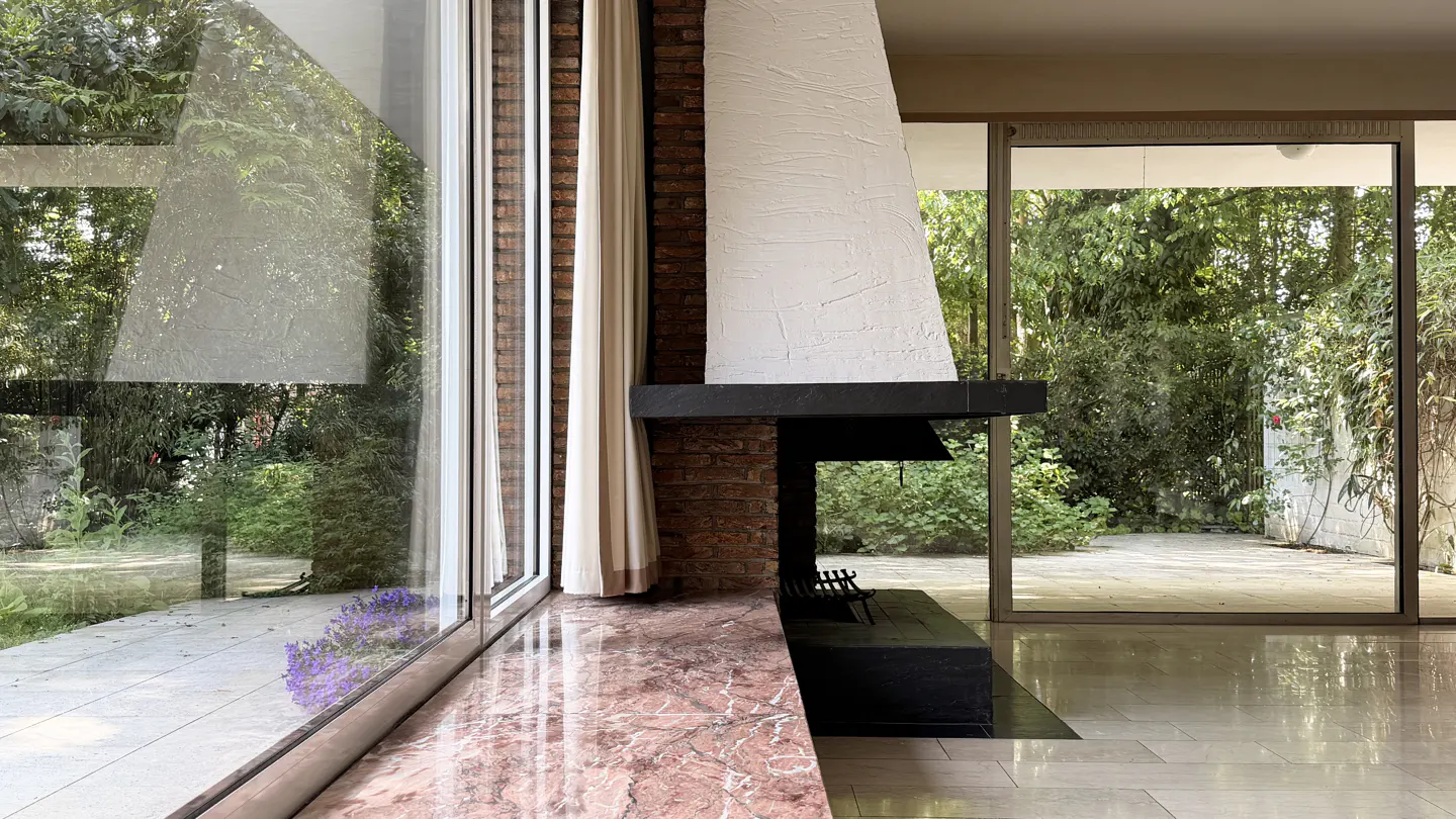 Interior view of a modern living room with a brick fireplace, large windows, and marble floors. Outside, lush greenery is visible.