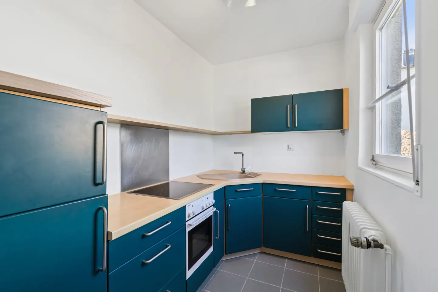 A compact kitchen with teal cabinets, light countertops, and white walls. A window is on the right, and a radiator is below it.