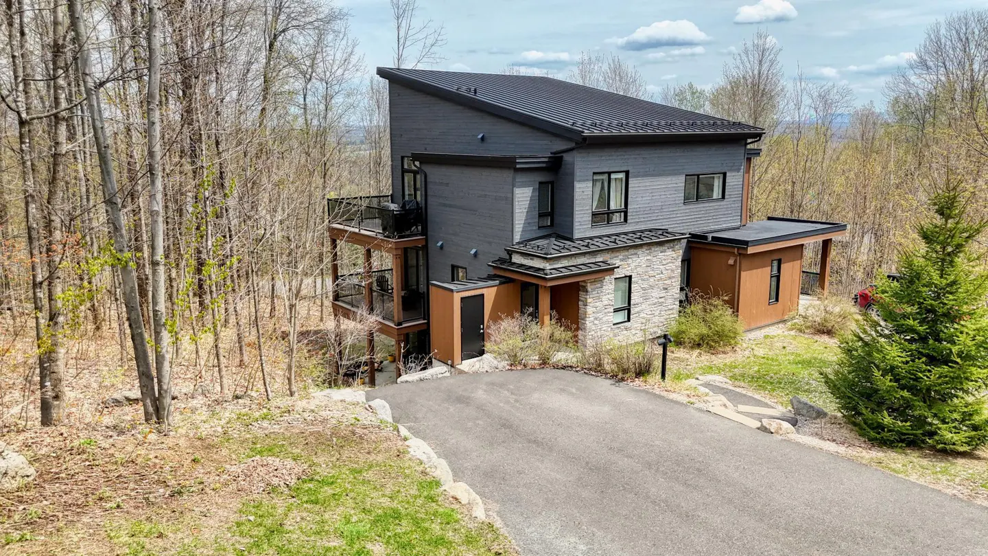 Modern gray house with black roof and stone accents, surrounded by trees and a paved driveway. Balconies are visible on the side.