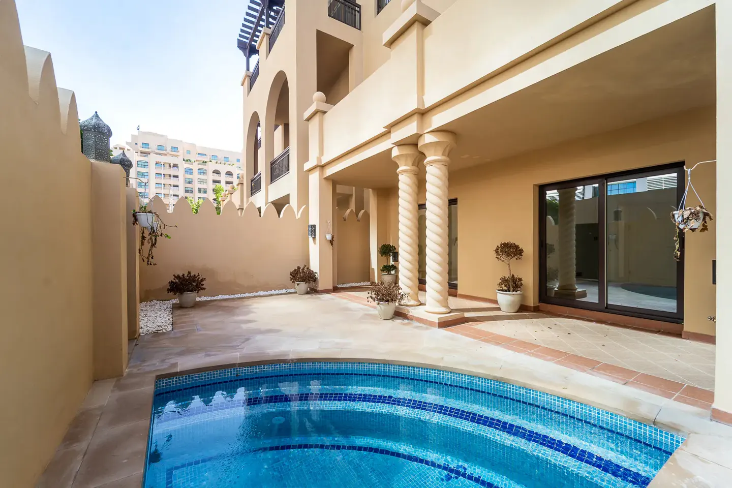 A tan villa with a blue tiled pool in the foreground. The villa has a covered patio with spiral columns. Potted plants are scattered around the patio.