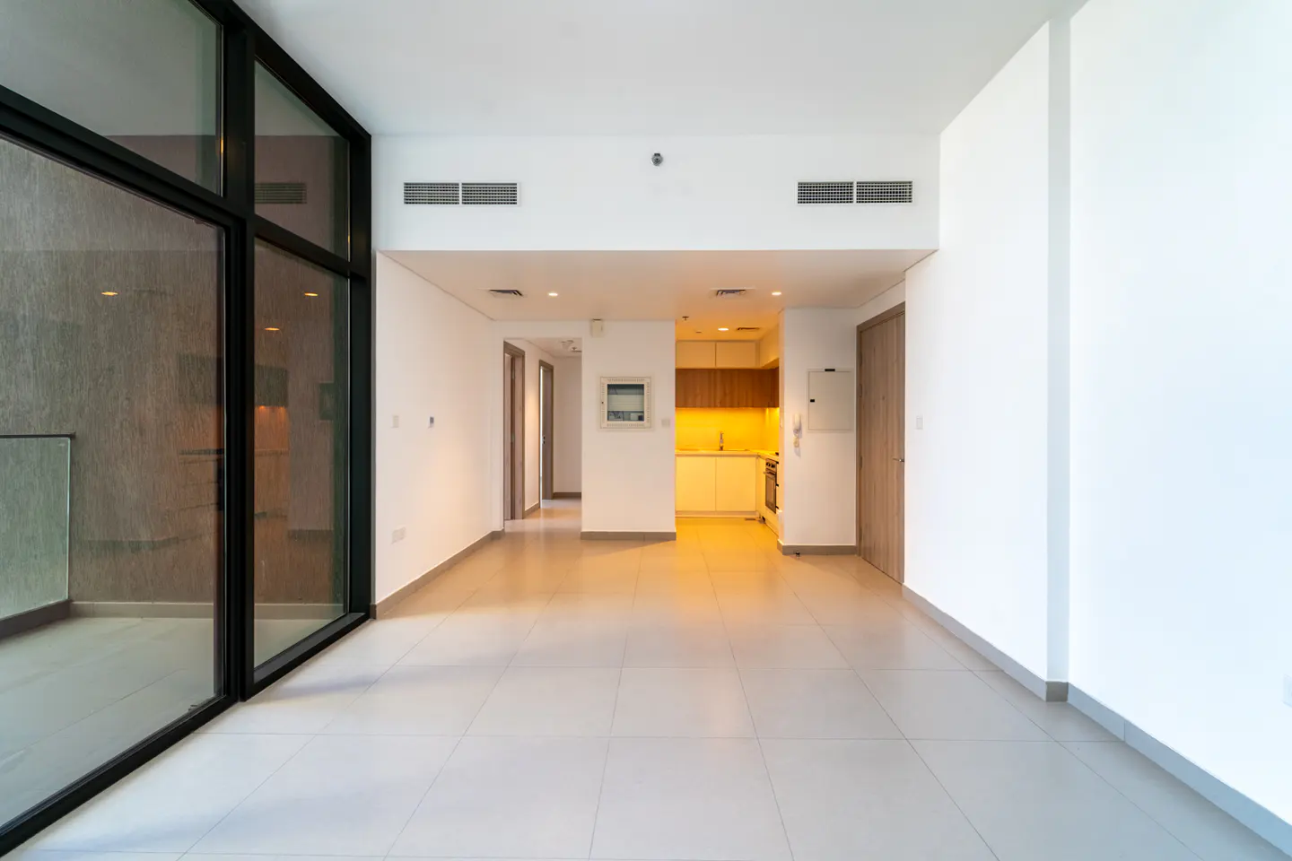 An empty, modern apartment with white walls, gray tile floors, and a black-framed window. A kitchen is visible in the background.