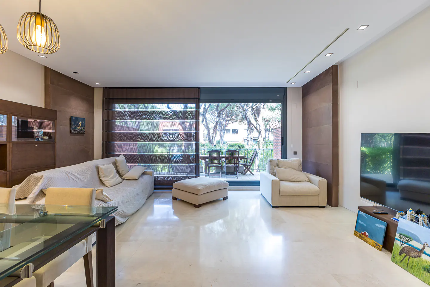 Bright living room with beige sofa, chair, and ottoman. A glass table sits in the foreground. Balcony with table and chairs visible through sliding glass doors.