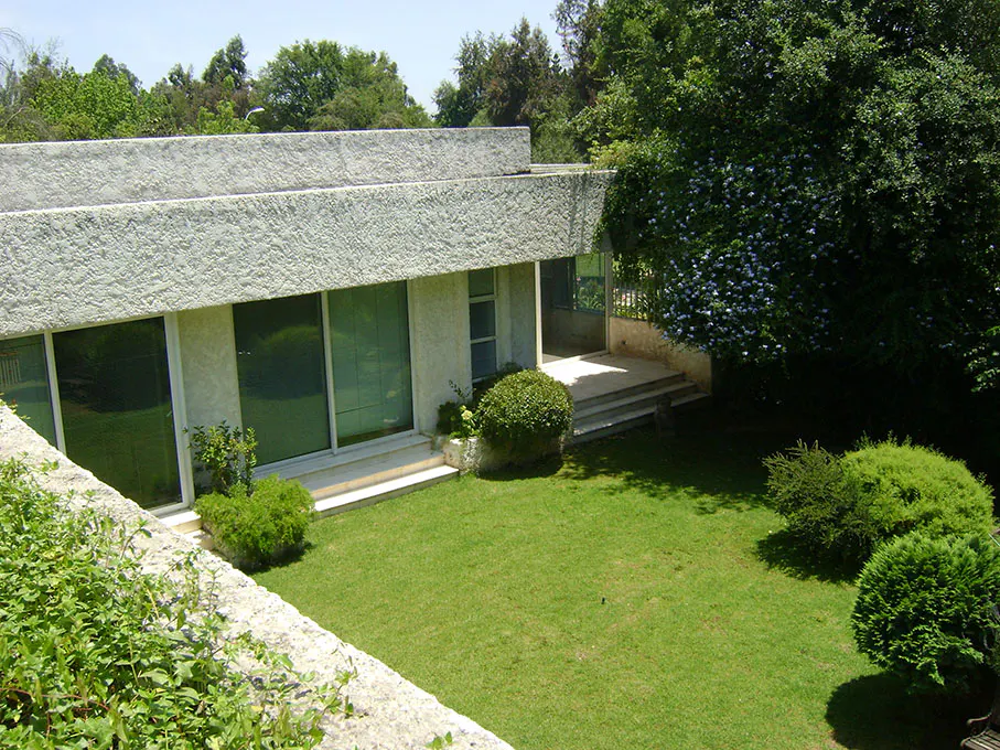 Exterior view of a modern, single-story house with a textured white facade, large windows, and a green lawn with bushes.