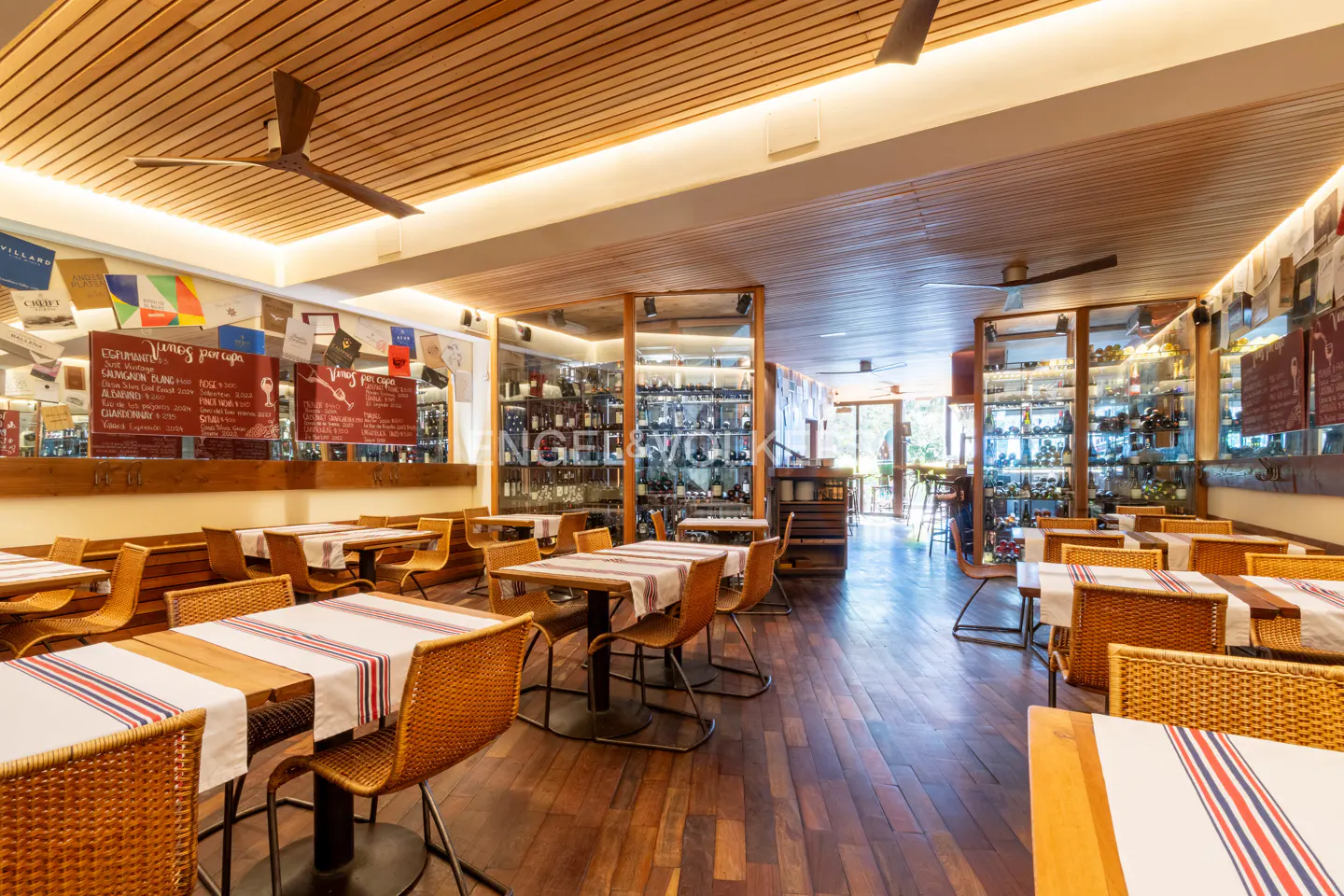 Interior of a restaurant with wooden floors, tables with striped runners, and wicker chairs. Wine bottles are displayed on shelves.