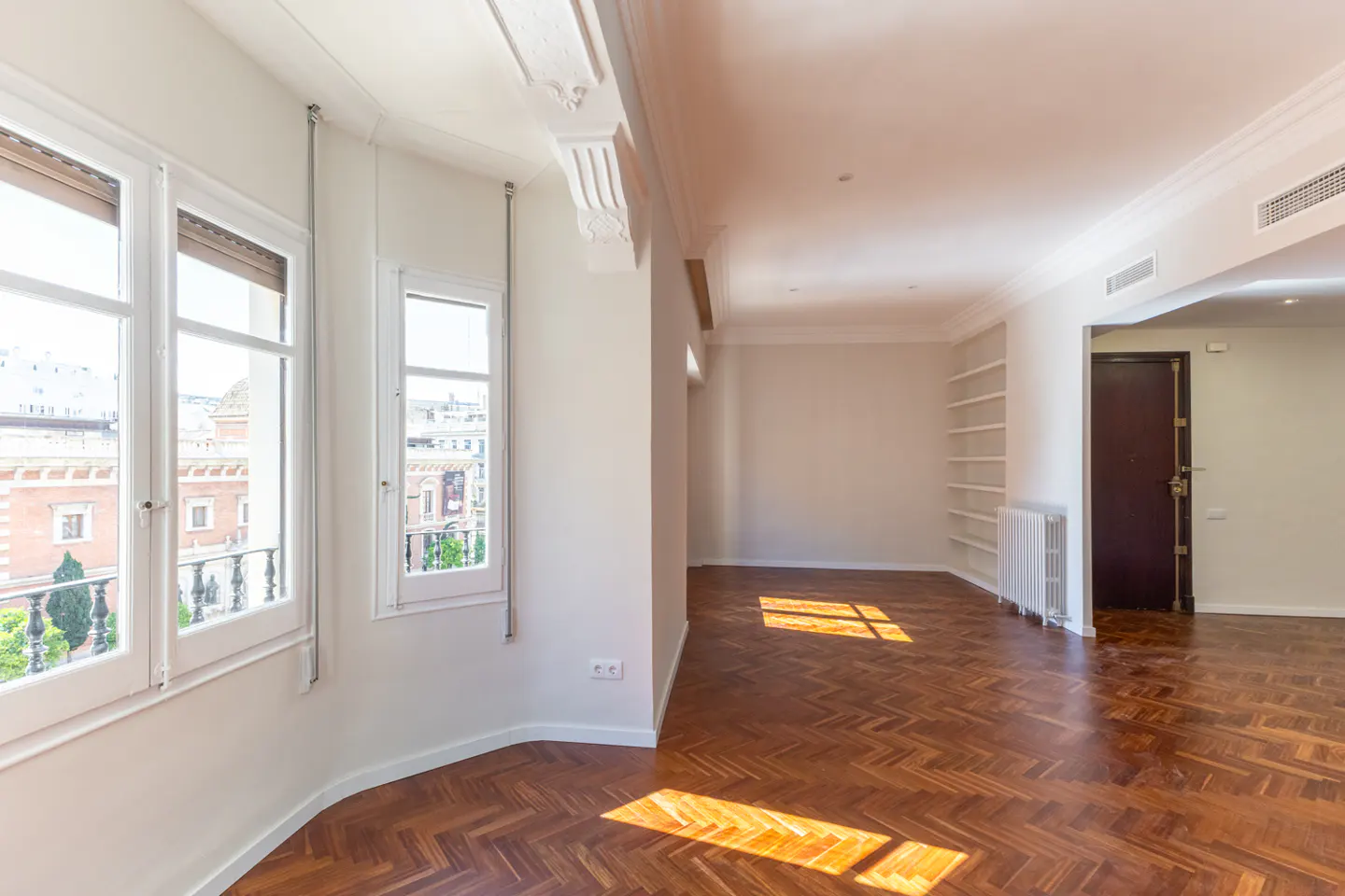 Bright, empty room with herringbone wood floors, white walls, and large windows overlooking a city.
