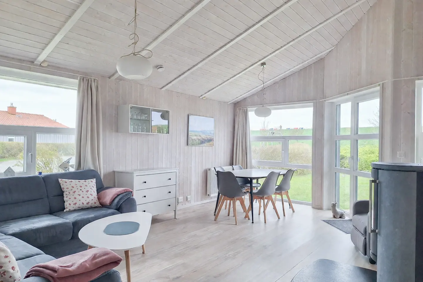 Bright living room with white wood paneling, blue sofa, dining table, and large windows overlooking a green landscape.