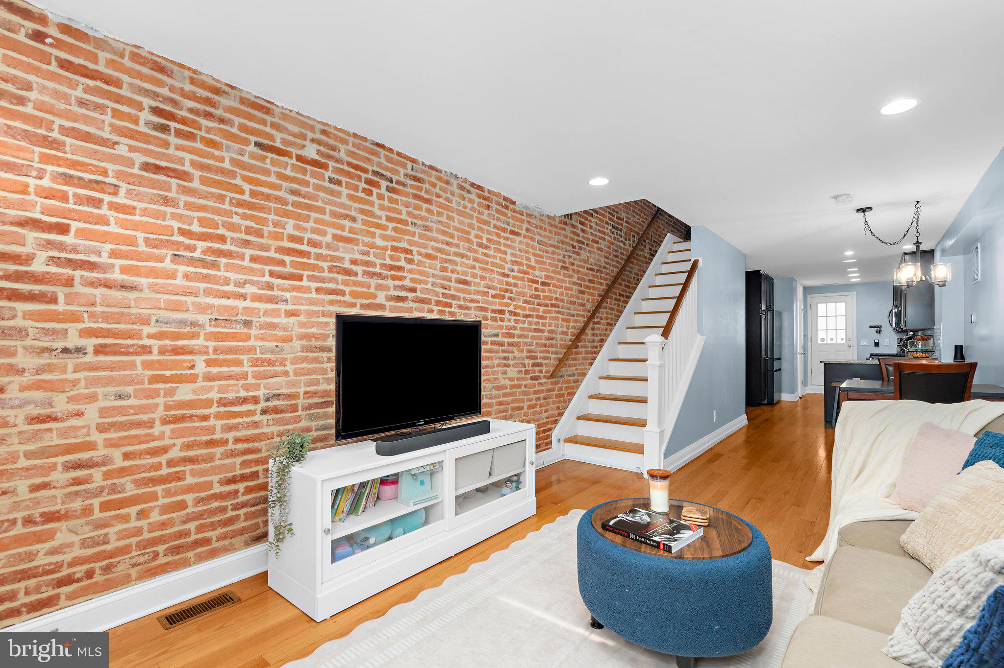 Living room with exposed brick wall, flat-screen TV on a white media console, wooden stairs, and a blue ottoman. [AI-generated text]