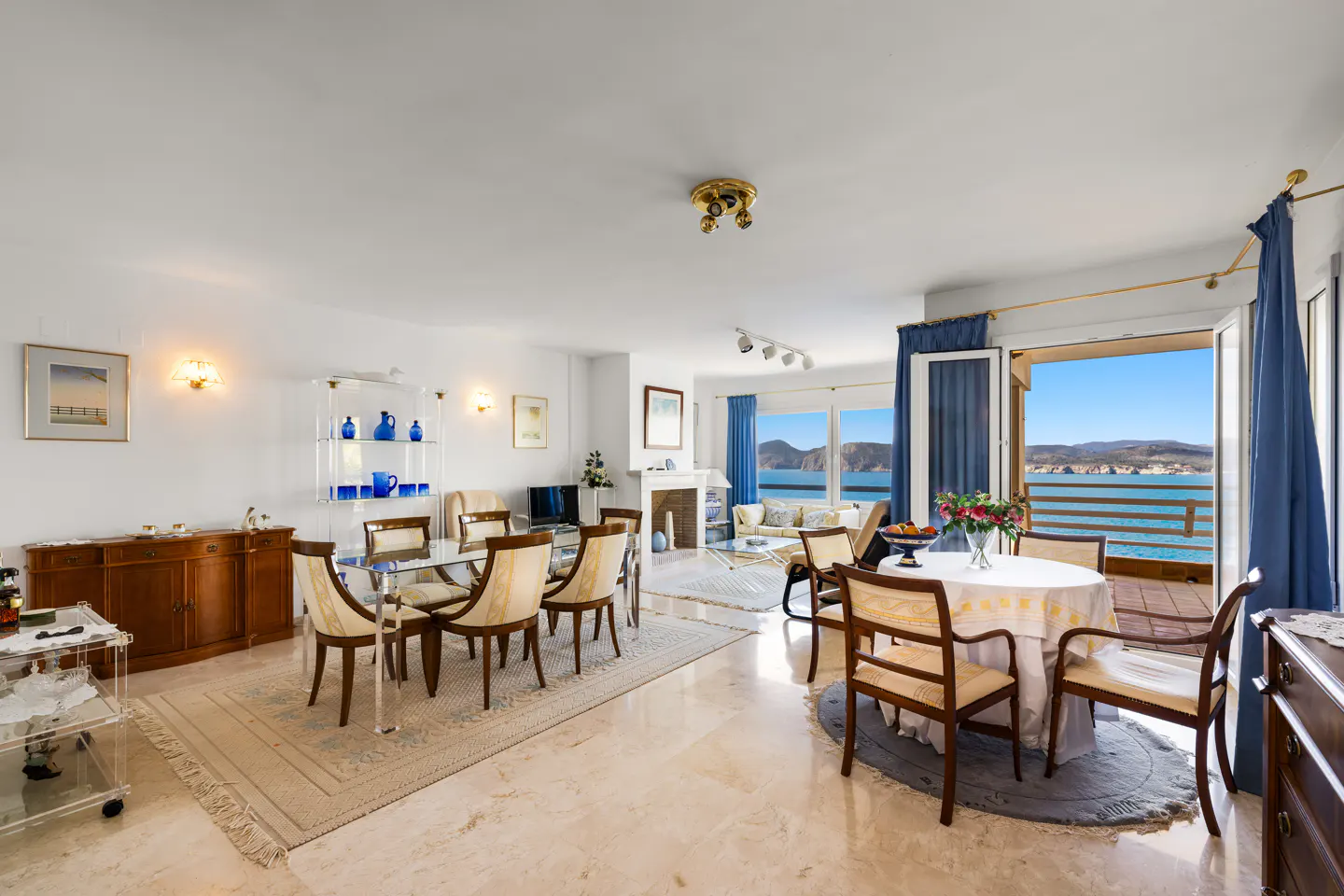 Bright, open-plan living and dining area with marble floors and ocean view. Blue curtains frame the balcony doors.
