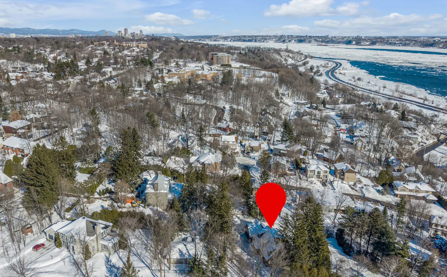 Aerial view of a snow-covered neighborhood with a red location pin on a house. A river and city skyline are in the background.
