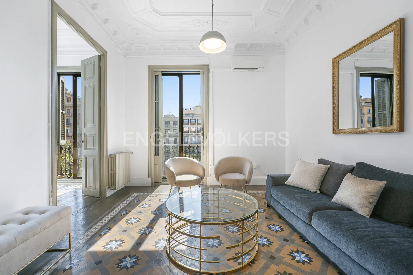 Bright living room with blue sofa, two beige chairs, and a glass coffee table on a patterned tile floor. A mirror hangs on the white wall.