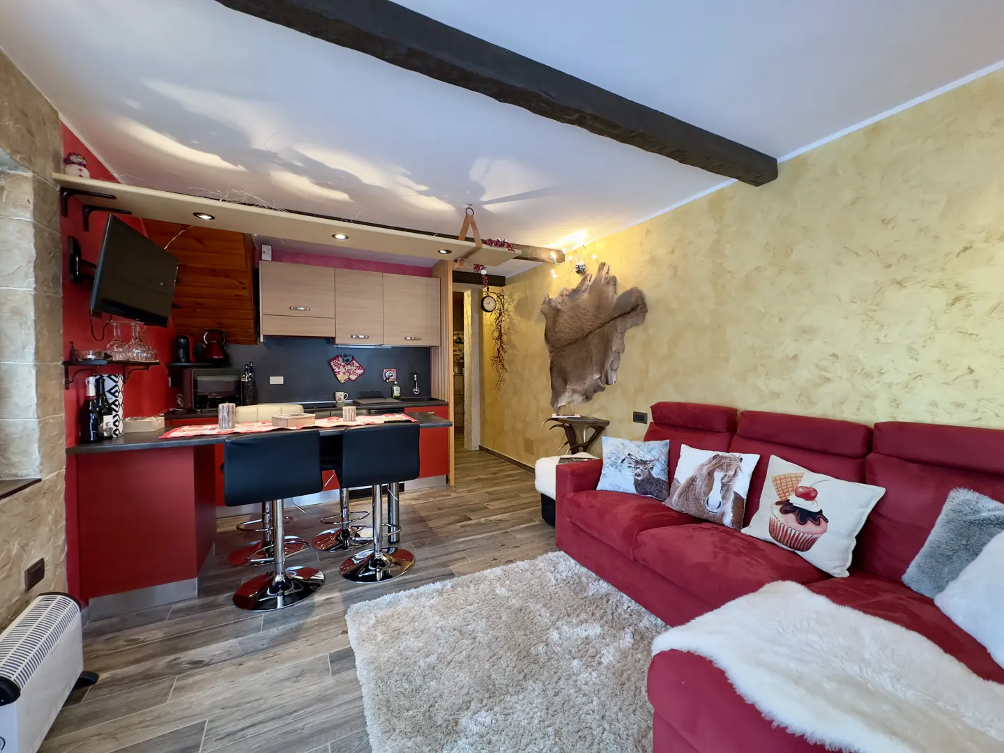 Cozy living space with a red sofa, animal-themed pillows, and a kitchen area with bar stools. A decorative animal hide hangs on the textured wall.