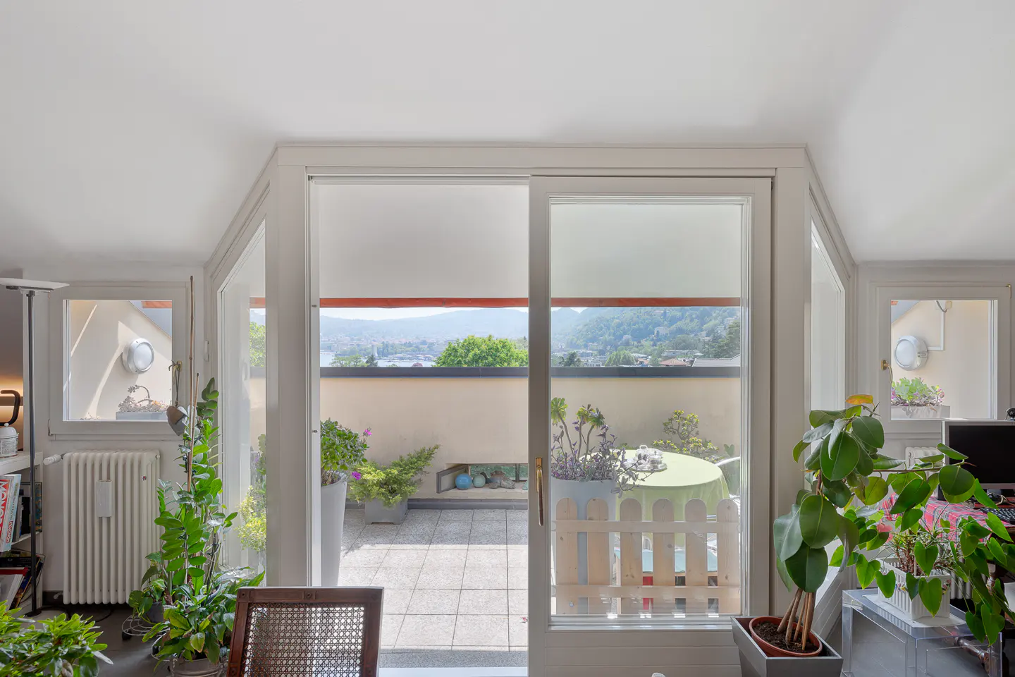 Bright room with open sliding doors to a balcony. Plants inside and outside. View of a lake and mountains.