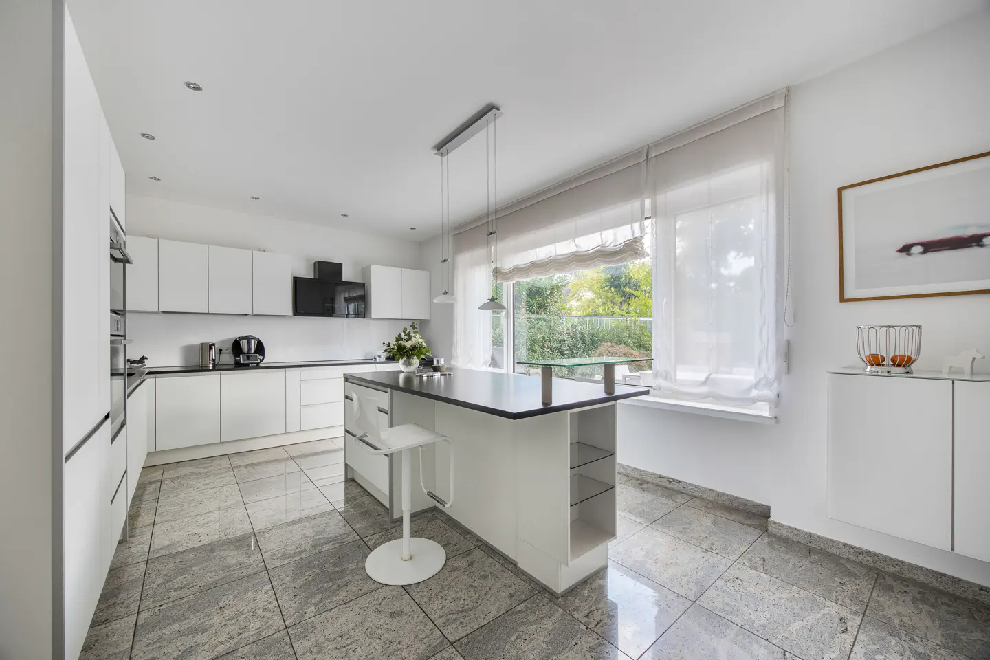Bright, modern kitchen with white cabinets, black countertops, and a gray granite floor. A central island has a black top and white base with a stool. Large window with sheer curtains.