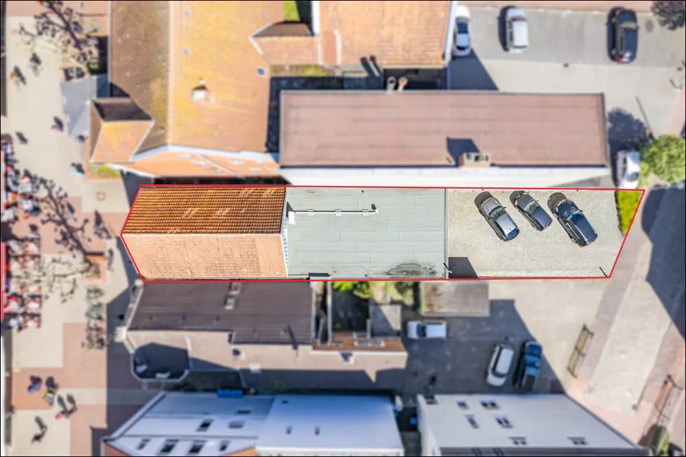 Aerial view of a building with a red-tiled roof and a flat roof section featuring three parked cars, outlined in red.