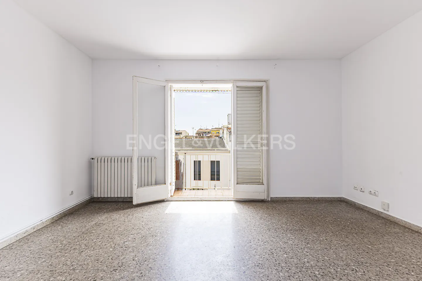 Bright, empty room with white walls and terrazzo floor. Open balcony doors reveal city view. Radiator on left wall.