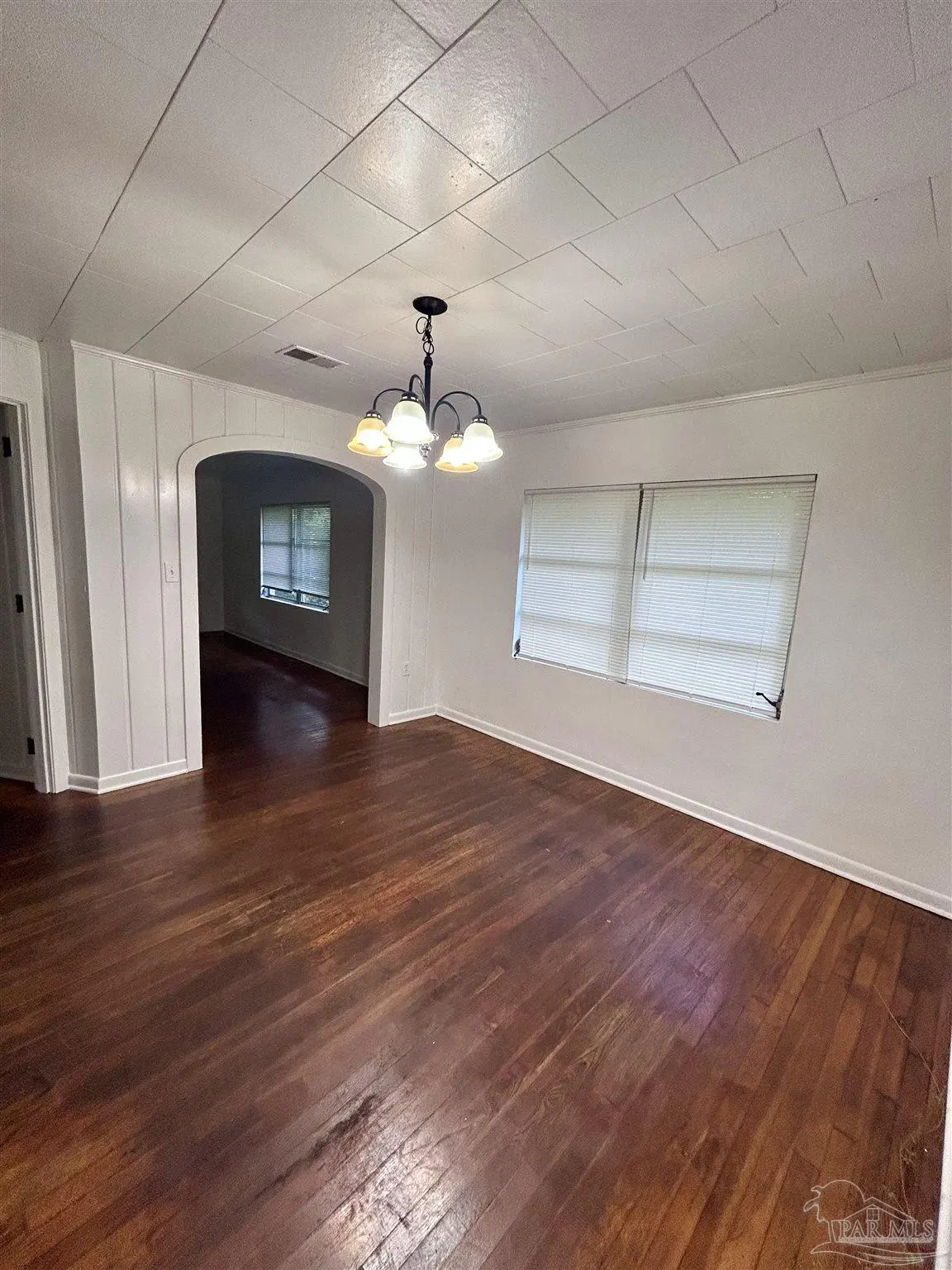 Empty dining room with dark wood floors, white walls, a chandelier, and an arched doorway leading to another room.